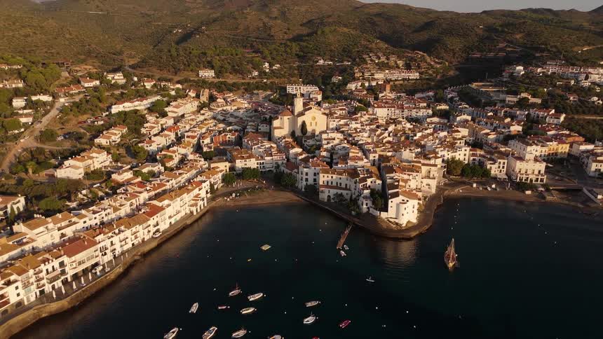 An aerial view captures a cluster of whitewashed buildings nestled between lush rolling hills and a deep blue baysmall boats dot the calm water in the foreground while a prominent church with a tall bell tower stands at the heart of the coastal tow