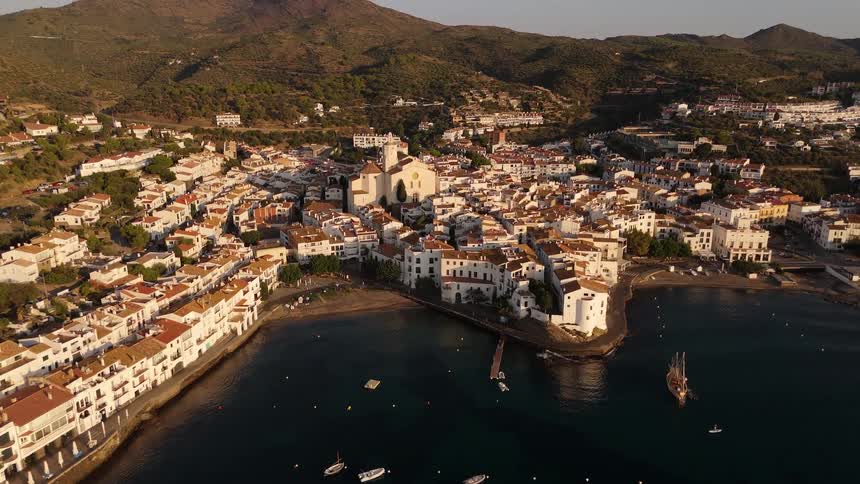 A coastal town with buildings lining the shoreline the orange setting sun casting a warm glow across the city with mountains rising in the background