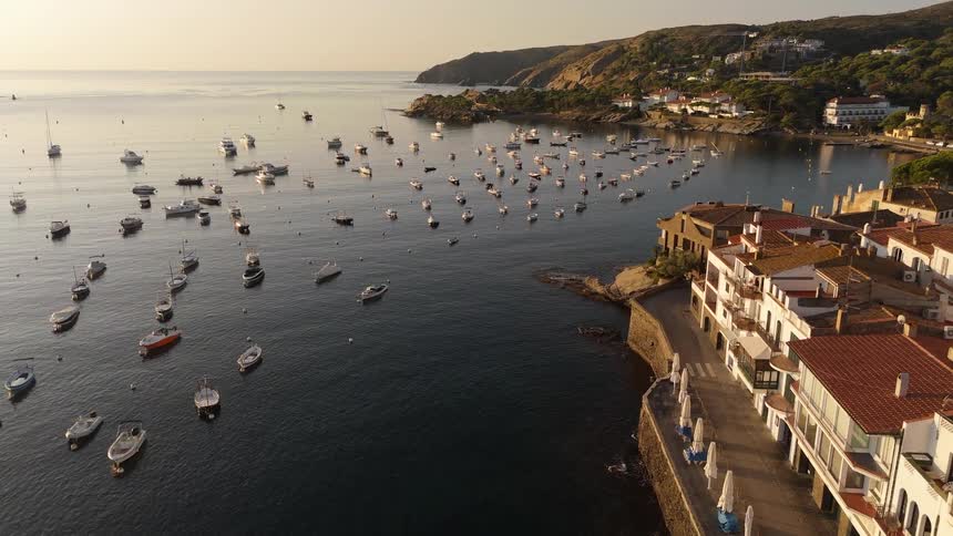 Aerial view of a calm coastal harbor filled with anchored boats beside a seaside village at sunset