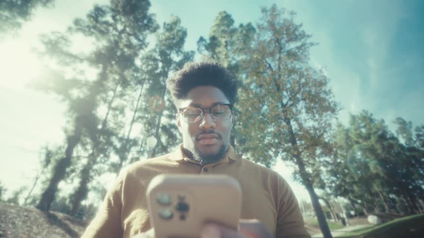 A man standing in an open space on a sunny day typing on a smartphone with trees behind him
