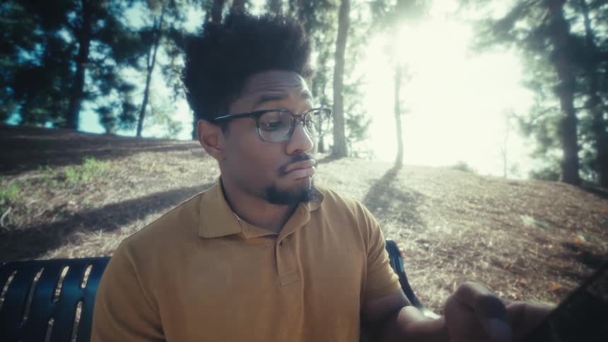 A man with glasses sits on a park bench and picks up his smartphone to check the screen while sunlight filters through the trees in the background