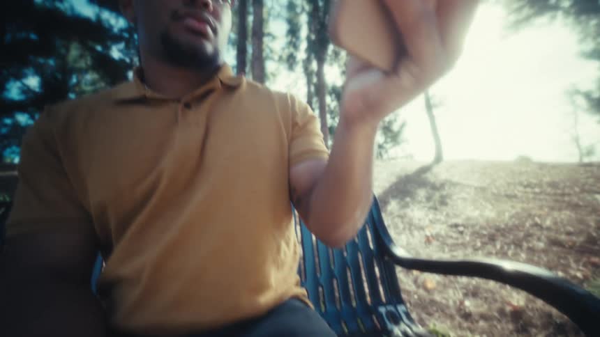 A young man wearing glasses sits on a bench in a public park at midday and picks up the smartphone that had been resting on his lap