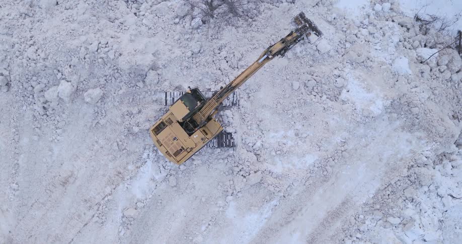 An aerial top down view shows a large yellow excavator using its long hydraulic arm and bucket to move and pile thick layers of white snow and icy debris across a wide frozen clearing surrounded by scattered bare trees and muddy tracks