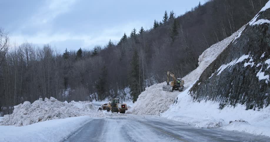 Several heavy machines clear a massive pile of snow that has blocked a road next to a steep mountain slope