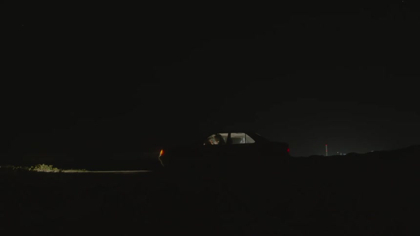 Person sits in a parked car at night as the headlights illuminate the grass