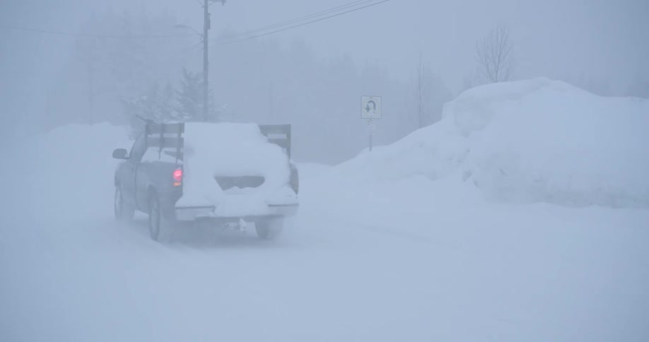 A dark pickup truck with a wooden rack drives through a heavy blizzard on a snow covered road past a uturn sign and buried embankments under a grey sky
