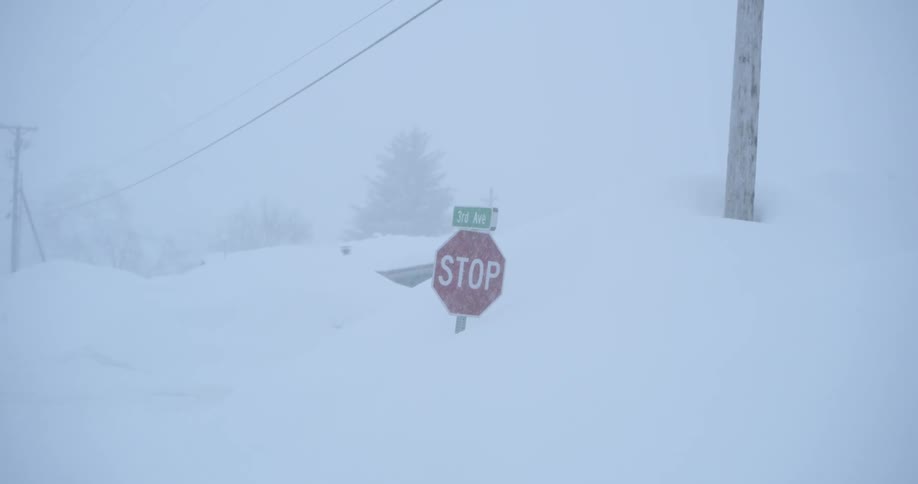 A stop sign and a 3rd ave street sign are almost completely buried by massive snowdrifts during a heavy blizzard as the camera zooms out a vehicle with glowing headlights slowly navigates through the whiteout conditions in the distance