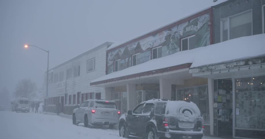 A snowy street in a small town features parked cars and buildings covered in snow during a winter storm