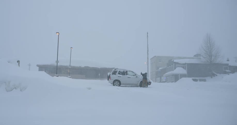A person shovels snow off a car on a rural street during a heavy blizzard