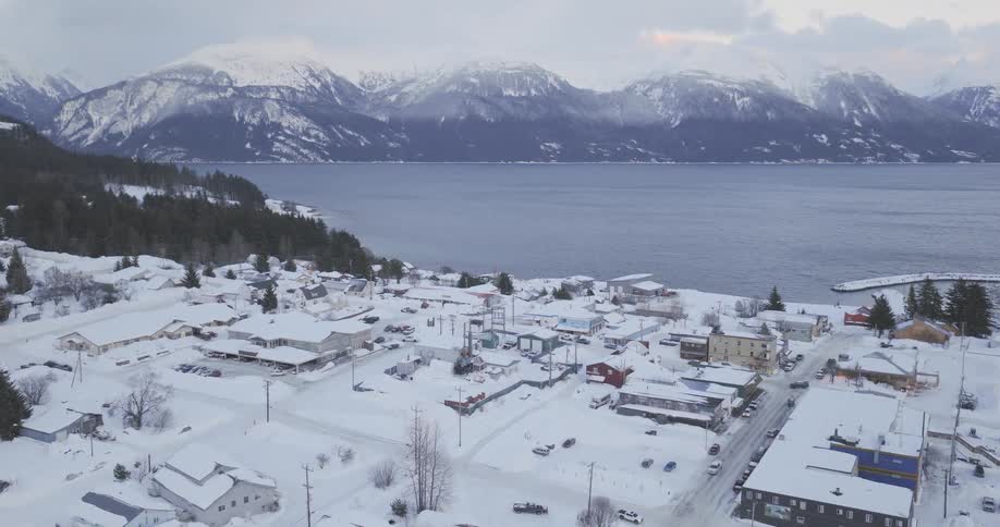 A drone captures a wide aerial view of a small coastal town covered in deep white snow nestled between a dark blue bay and a massive range of jagged snow capped mountains under a gloomy winter sky