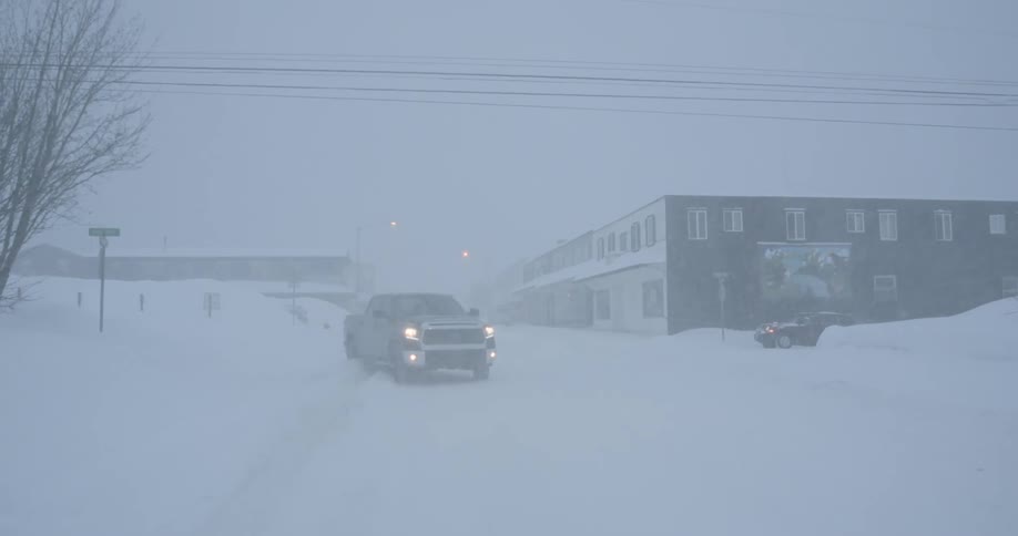 A dark pickup truck carefully turns onto a deserted town street its headlights cutting through the heavy blowing snow of an intense blizzard that blankets the entire landscape