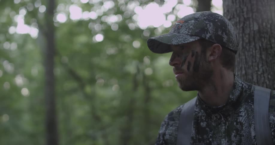 A hunter is searching the forest as he sits in a tree