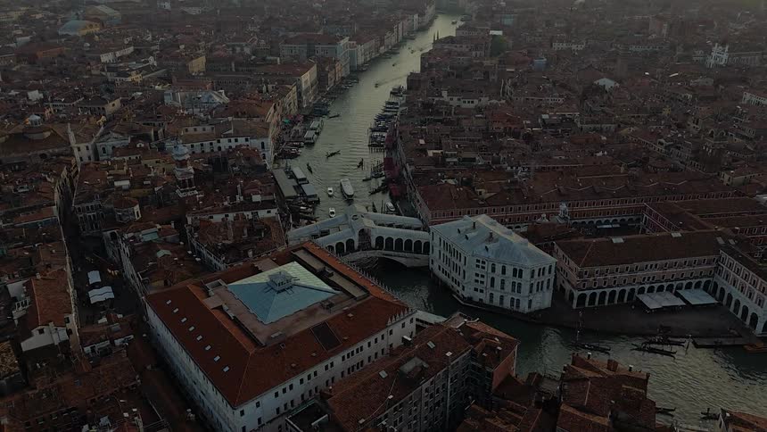 An aerial shot captures the densely packed terracotta rooftops and the bustling grand canal of venice with the rialto bridge at its center