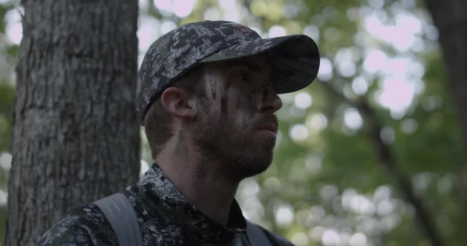 A man dressed in camouflage clothing is leaning against a tree looking around intently