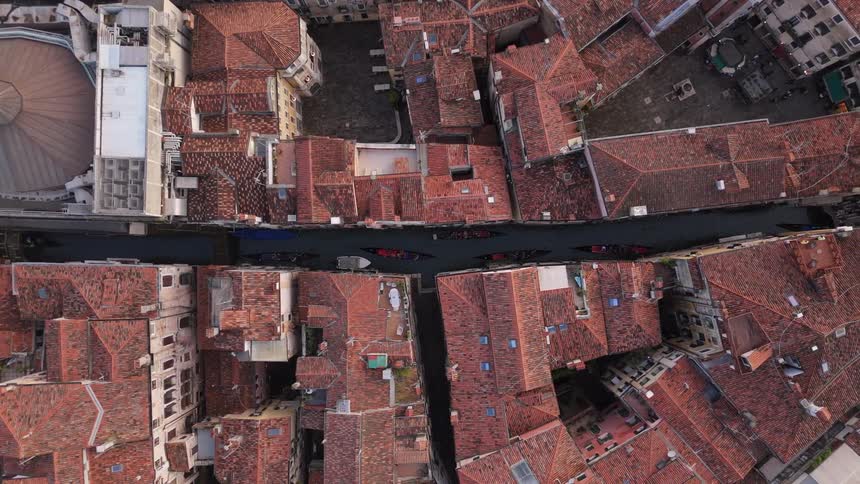 An aerial view shows several gondolas moving through a narrow canal that cuts between old buildings with red tiled roofs in a city