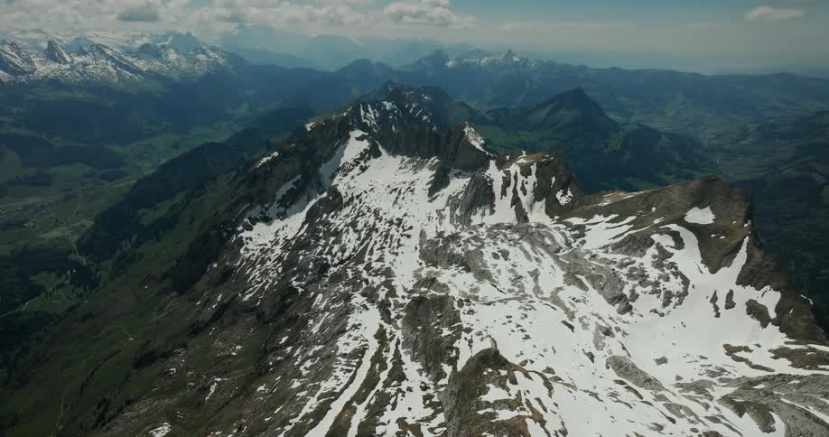 An aerial view captures a jagged mountain ridge partially covered in patches of white snowlush green valleys and distant hazy mountain ranges stretch toward the horizon under a cloudy sky