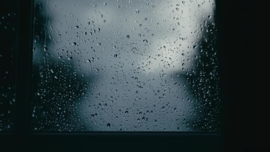 Raindrops cling to and slowly trickle down a windowpane blurring the view of a gloomy overcast world outside into a moody abstract pattern