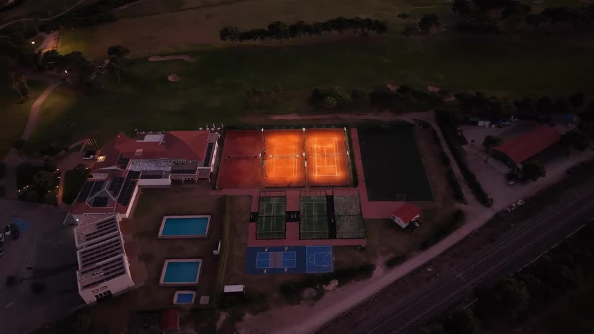 An aerial view captures a sports complex at twilight featuring two brightly lit clay tennis courts that stand out against the darkening landscape the facility includes various athletic areas like swimming pools and basketball courts all situated next t