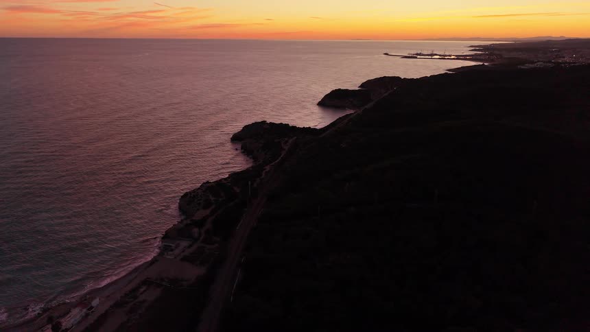 An aerial view shows a dark coastline at sunset where the ocean meets a rocky shore and a forest
