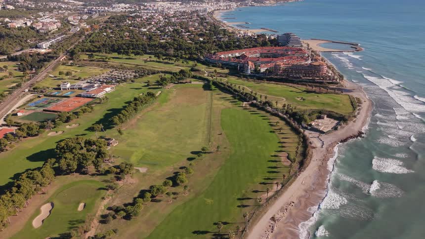 A high angle drone shot moves over a green golf course and a sandy beach where turquoise waves crash on the shore near a large red roofed resort complex and a sprawling coastal city