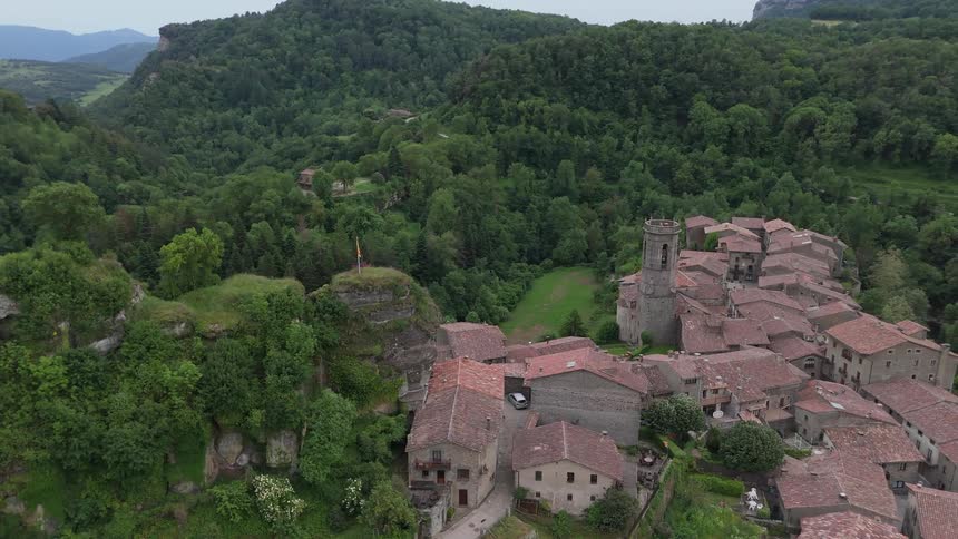 An aerial view shows an old stone village with many brown tile roofs nestled in a green valley surrounded by lush forests and mountains