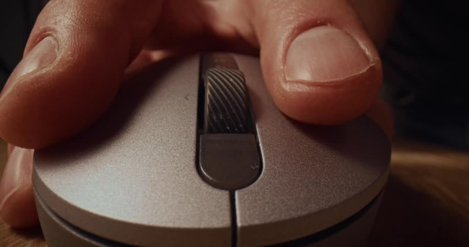 Fingers grip and operate the scroll wheel of a silver computer mouse on a wooden desk