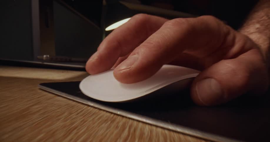 A hand clicks a white computer mouse on a wooden desk in a dimly lit workspace