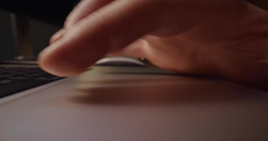 A close up shot shows a person using their index and middle fingers to tap and scroll on a laptop trackpad in a dimly lit room
