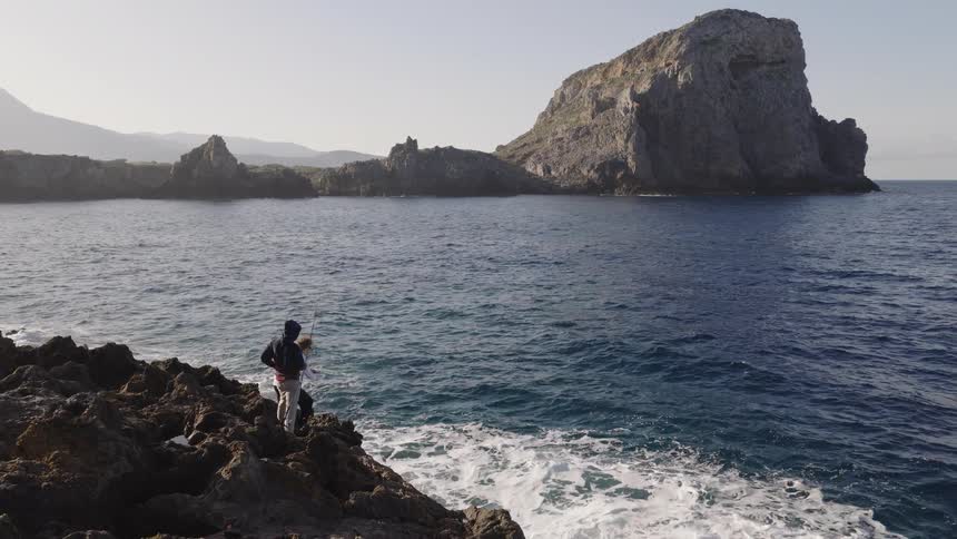 A person in a dark jacket stands on a rugged rocky coastline looking out toward a massive rock formation rising from the seawaves crash against the jagged shore in the foreground while the camera slowly pulls back to reveal the scale of the coastal