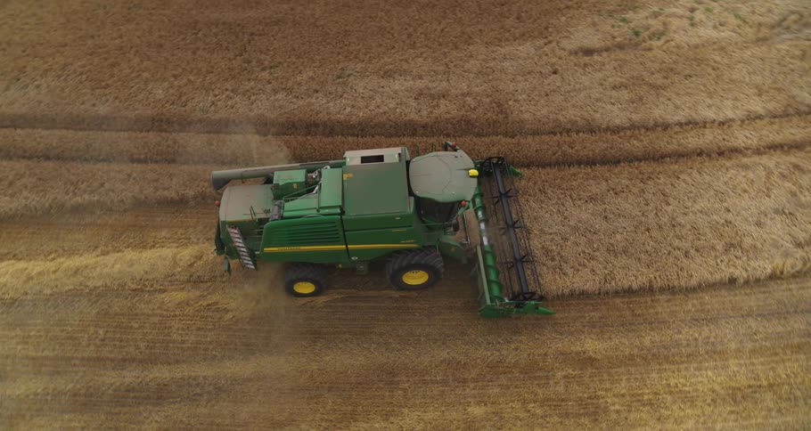 An aerial view shows a green combine harvester actively working through a large golden brown field