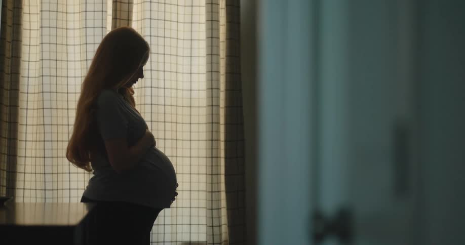 Pregnant woman in profile with hands on her belly in front of a lit curtain