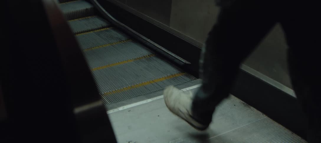 A person wearing dark pants and white shoes steps onto a moving escalator in a dimly lit area