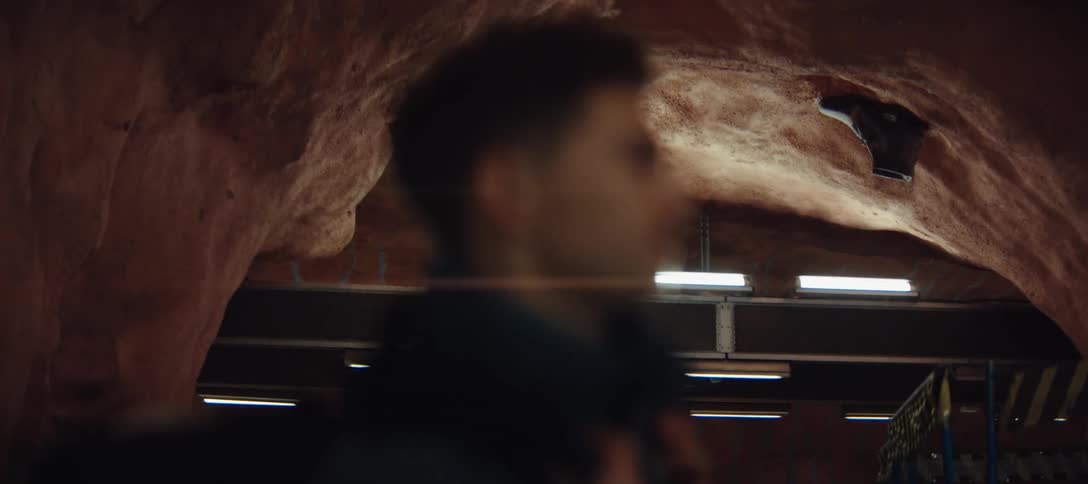 A man with short dark hair walks through a subway station built into a natural cave with textured red rock ceilings and modern industrial lighting