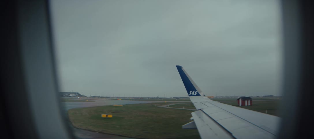 An sas airplane wing with a blue wingtip sits stationary on a damp runway under a heavy overcast gray skythe view from the cabin window captures a gloomy day at the airport showing green airfield grass and distant taxiway lights