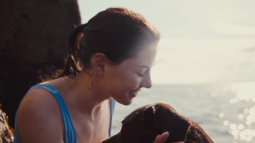 A smiling woman in a blue swimsuit sits by the ocean and pets a chocolate labrador as the sun shines behind them
