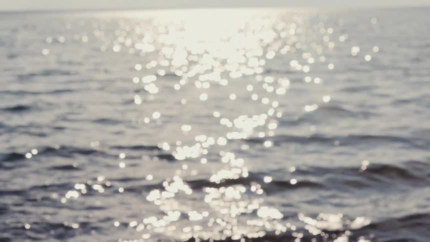 Sunlight reflects off the surface of the ocean waves as the camera slowly brings the rippling water into sharp focus during a bright day at the beach