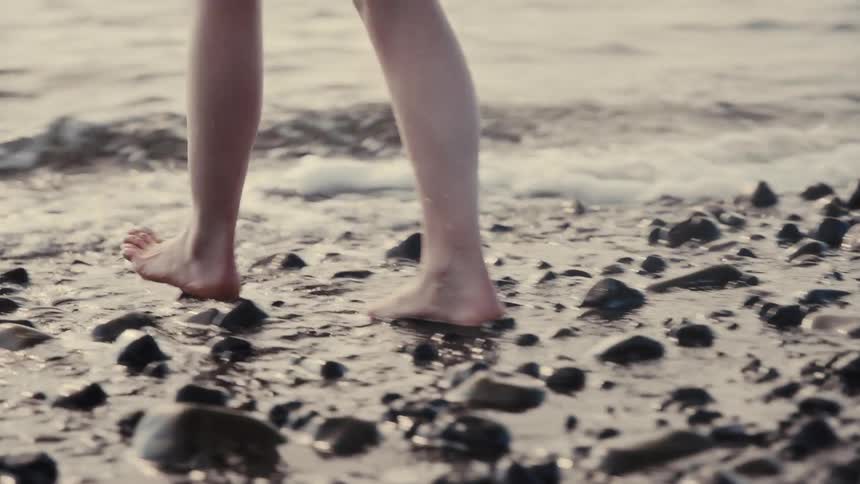 Woman walking barefoot on the coast and into the water as waves roll in to shore