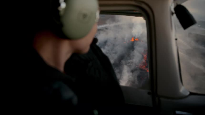 A woman wearing green headphones sits in the cockpit of a small airplane and looks out the side window at a volcanic eruption with red lava and white smoke below