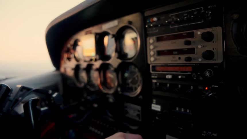 A pilots hand reaches out to adjust a knob on the complex instrument panel of a small aircraft the cockpit is bathed in the soft low light of dusk as the plane flies toward the glowing horizon