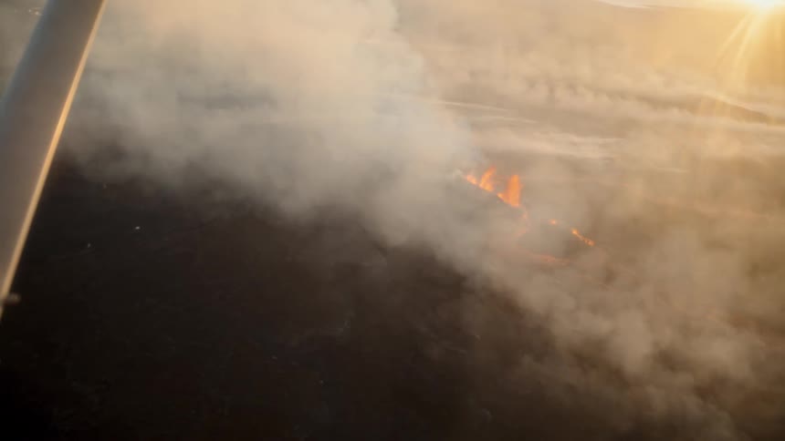 A fire burns across an open field at sunset sending up thick smoke that drifts into the sky and clouds the atmosphere