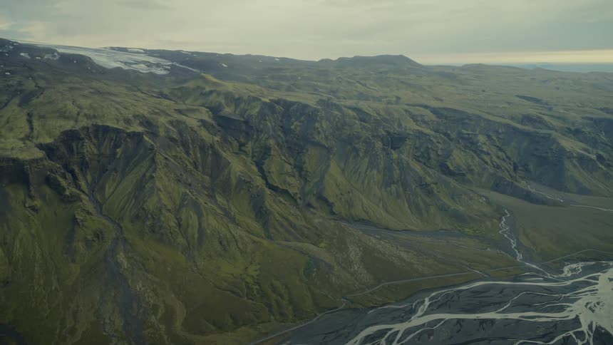 A highland landscape stretches out under a cloudy sky with a narrow stream running beneath the rocky ridge cutting gently through the terrain as the light stays soft and muted