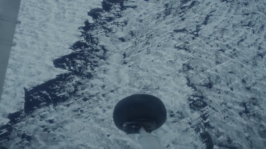 Landing gear visible flying over snowcovered terrain with exposed dark patches