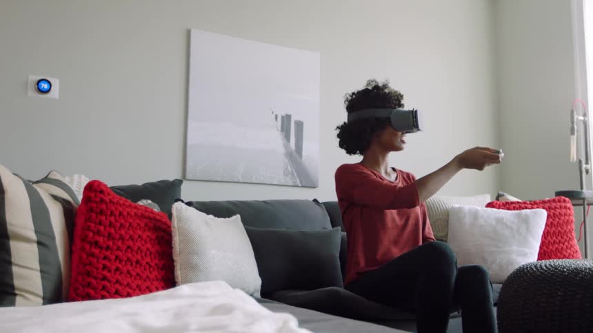 A woman wearing vr glasses sits on a couch and moves her arm