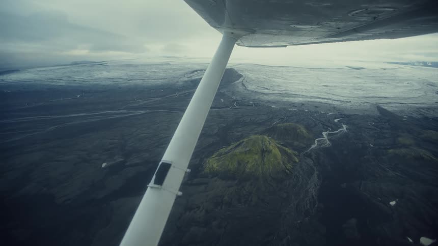 Aircraft wing flying over volcanic terrain with sparse vegetation and glacier in the distance under cloudy sky