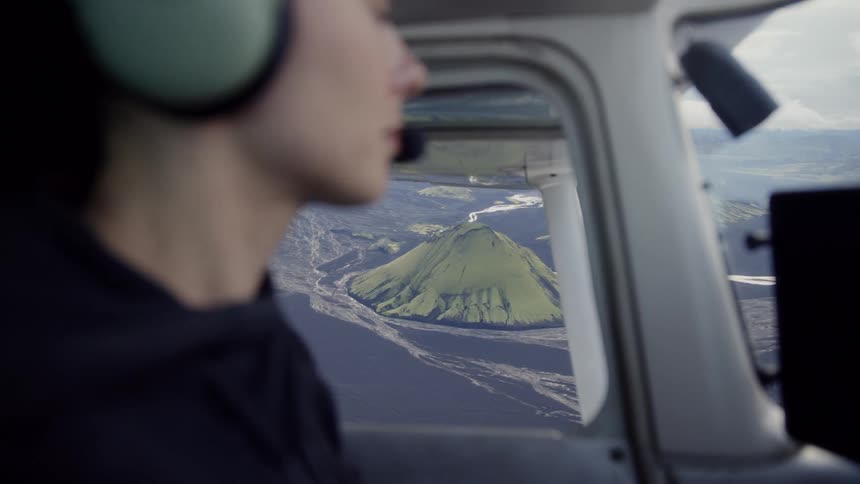 A person wearing a headset sits in the cockpit of a small aircraft and looks out the window at a vibrant green volcanic mountain surrounded by black sand and winding river veins in the icelandic highlands during a scenic flight