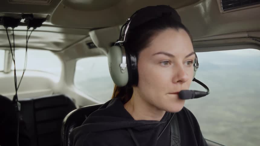 A woman wearing a headset talks into her microphone and gazes out the window while sitting in an airplane cockpit