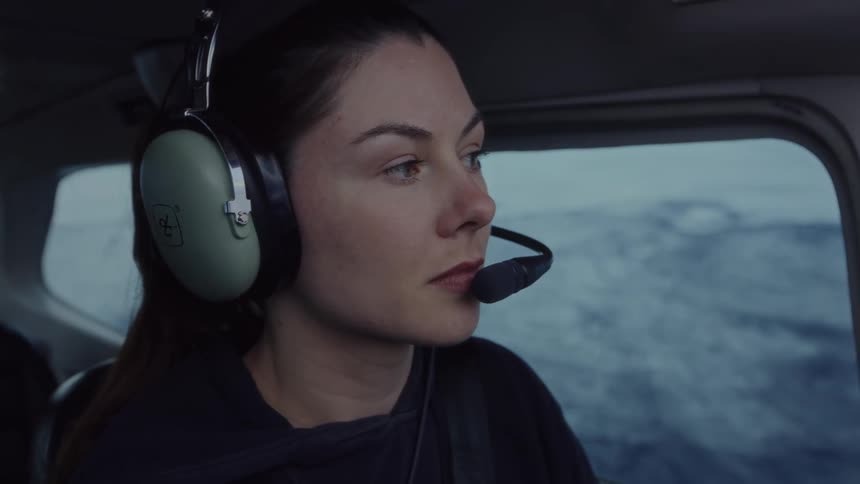 A woman with dark hair wearing green aviation headphones and a headset looks out the window of a small airplane while flying over a vast and snowy mountain landscape