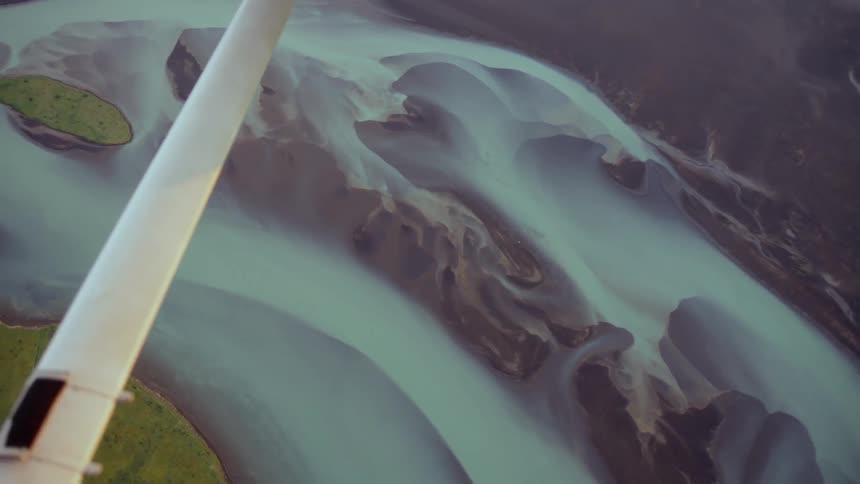 An aerial perspective from a small plane shows the intricate patterns of a glacial braided river flowing through dark silt and green coastal plains toward the sea