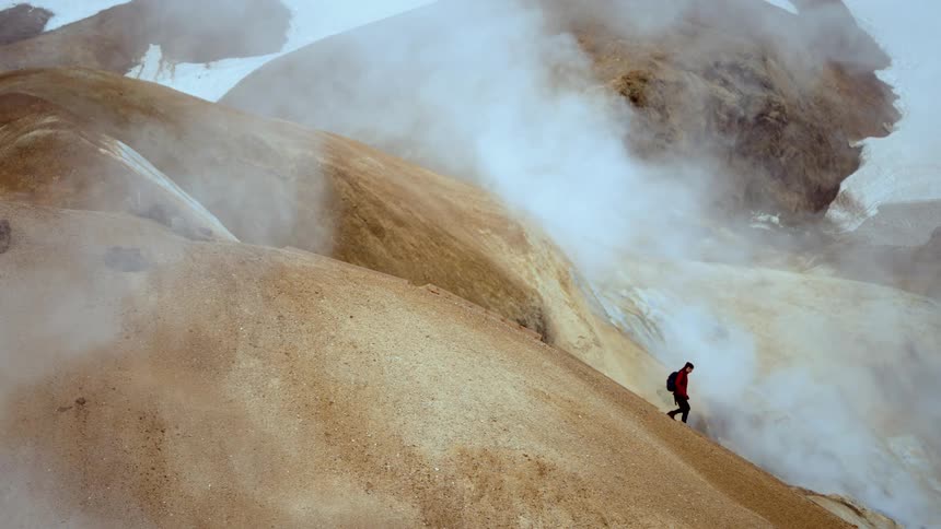 A person hikes down a volcanic mountain at midday smoke rising into the sky and creating a foggy atmosphere they walk steadily with a backpack on their back