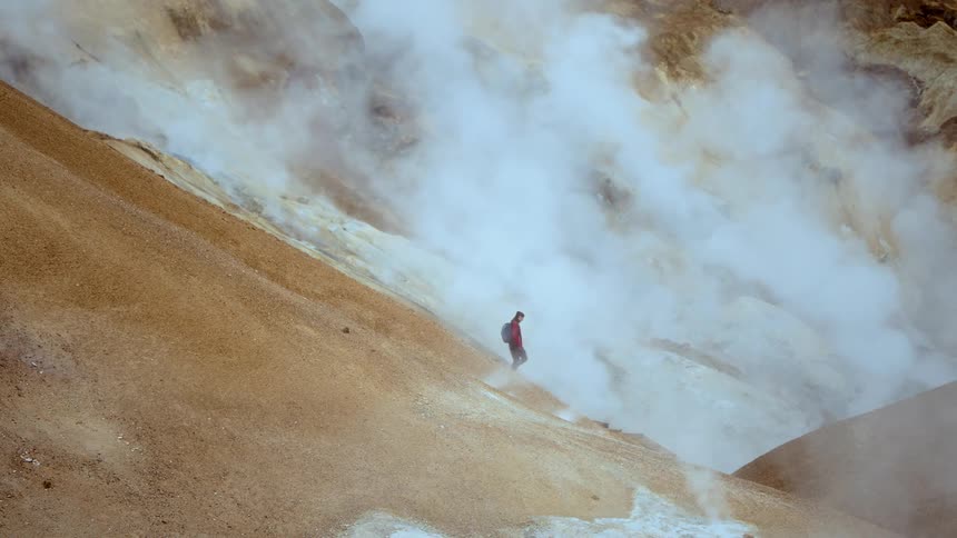 Person in red jacket walking on steep rocky terrain with geothermal steam rising around